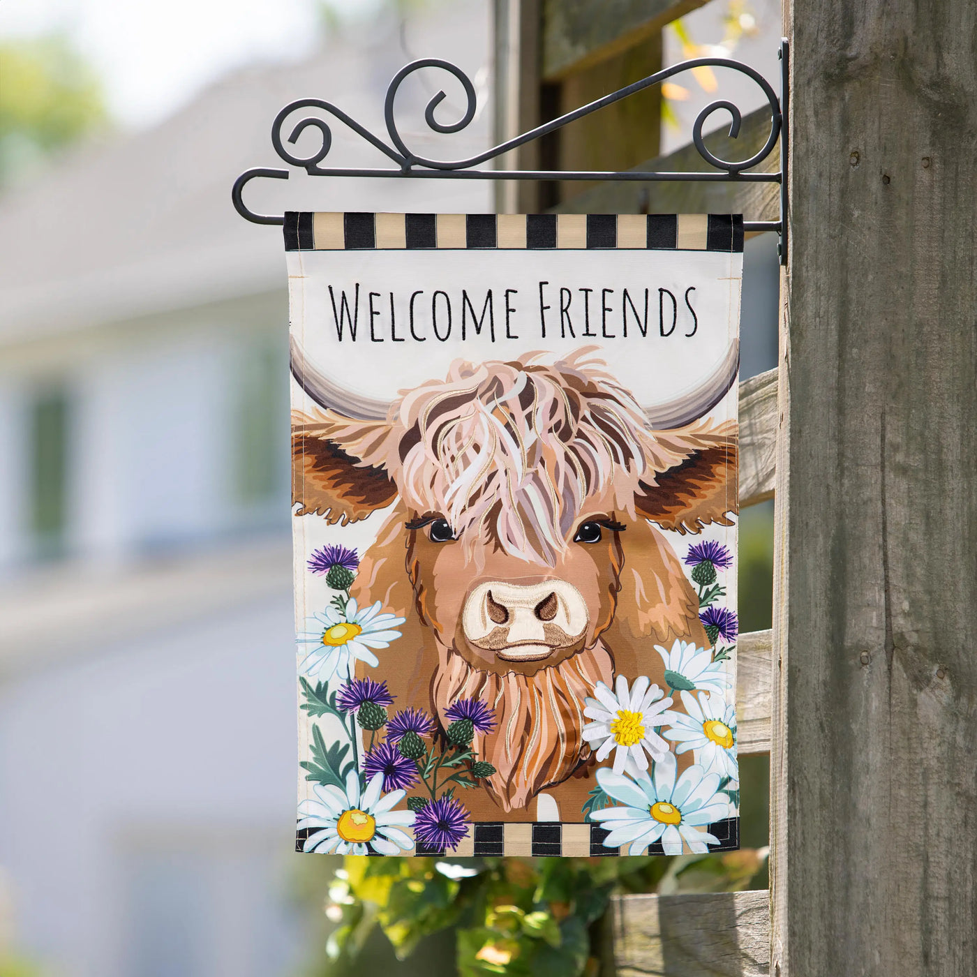 A cream hillside pasture garden flag with a brown highland cow applique surrounded by daisies and thistles